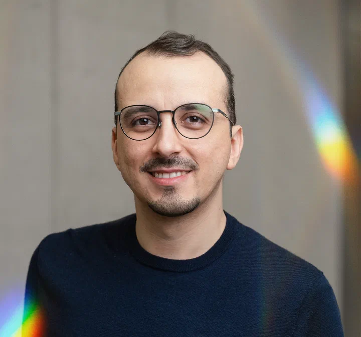 Italo Vietro smiling broadly, in glasses and a dark sweater, with a prism streak of light on the wall behind him.