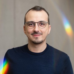 Italo Vietro, in glasses and a dark sweater, with a prism streak of light on the wall behind him.