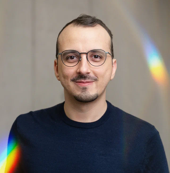 Italo Vietro, in glasses and a dark sweater, with a prism streak of light on the wall behind him.