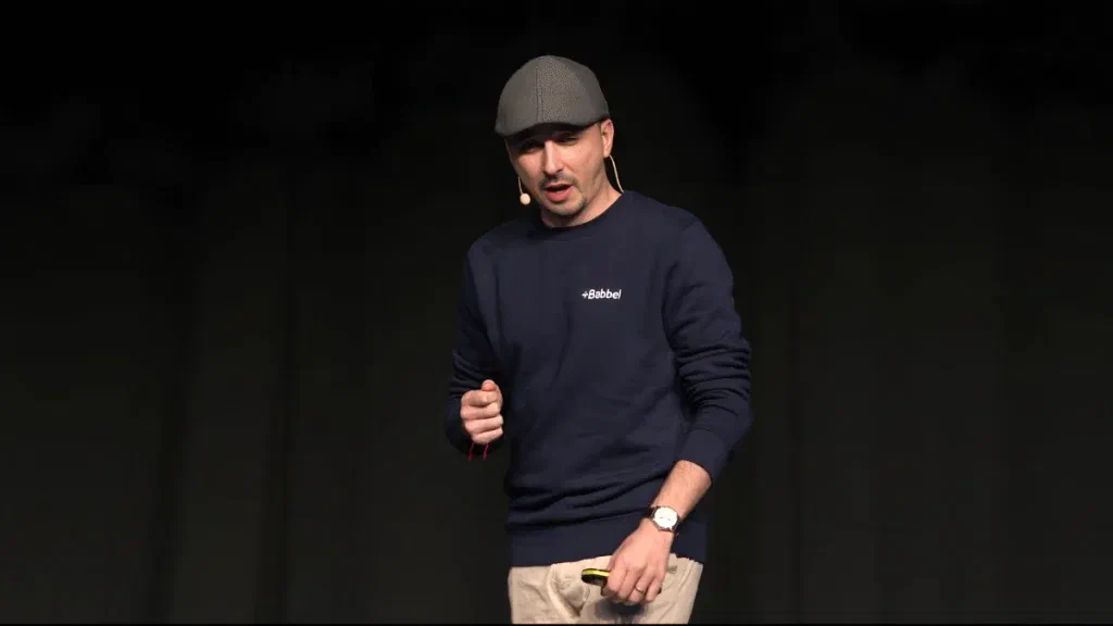 Italo Vietro on stage in a flat cap and a dark sweatshirt, speaking into a headset microphone against a dark backdrop.